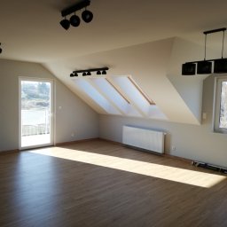 AKABUD - Poddasze po remoncie z drewnopodobną podłogą, trzema oknami dachowymi, grzejnikiem, lampami sufitowymi i listwami przypodłogowymi. Na podłodze leżą opakowania po panelach podłogowych.