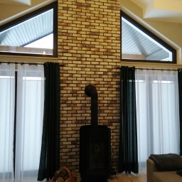 AKABUD - Salon z kominkiem opalanym drewnem, ściana z dekoracyjnej cegły, okna trójkątne, drewniana podłoga i białe zasłony.