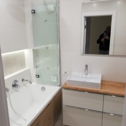 AKABUD - Nowoczesna łazienka z białą armaturą, kabiną prysznicową ze składanym szklanym panelem, umywalką nablatową na szafce z drewnianym blatem i lustrem, w którym odbija się osoba robiąca zdjęcie.