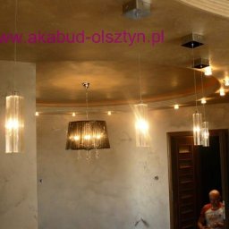 AKABUD - Wykończone wnętrze z dekoracyjną, metalizowaną ścianą i sufitem, oświetlone wiszącymi lampami z kryształkami, widoczny fragment otwartych drzwi i sylwetka mężczyzny.