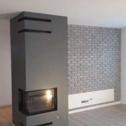AKABUD - Nowoczesny kominek obudowany szarą konstrukcją z poziomymi wnękami, z palącym się ogniem wewnątrz, na tle ściany wyłożonej szarymi cegłami w nowym wnętrzu z drewnianą podłogą.