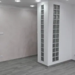 Wnętrze po remoncie: szare panele podłogowe imitujące drewno, ściany w odcieniach bieli i beżu, kolumna z luksferami, szara, nowoczesna stolarka drzwiowa z poziomymi wstawkami.