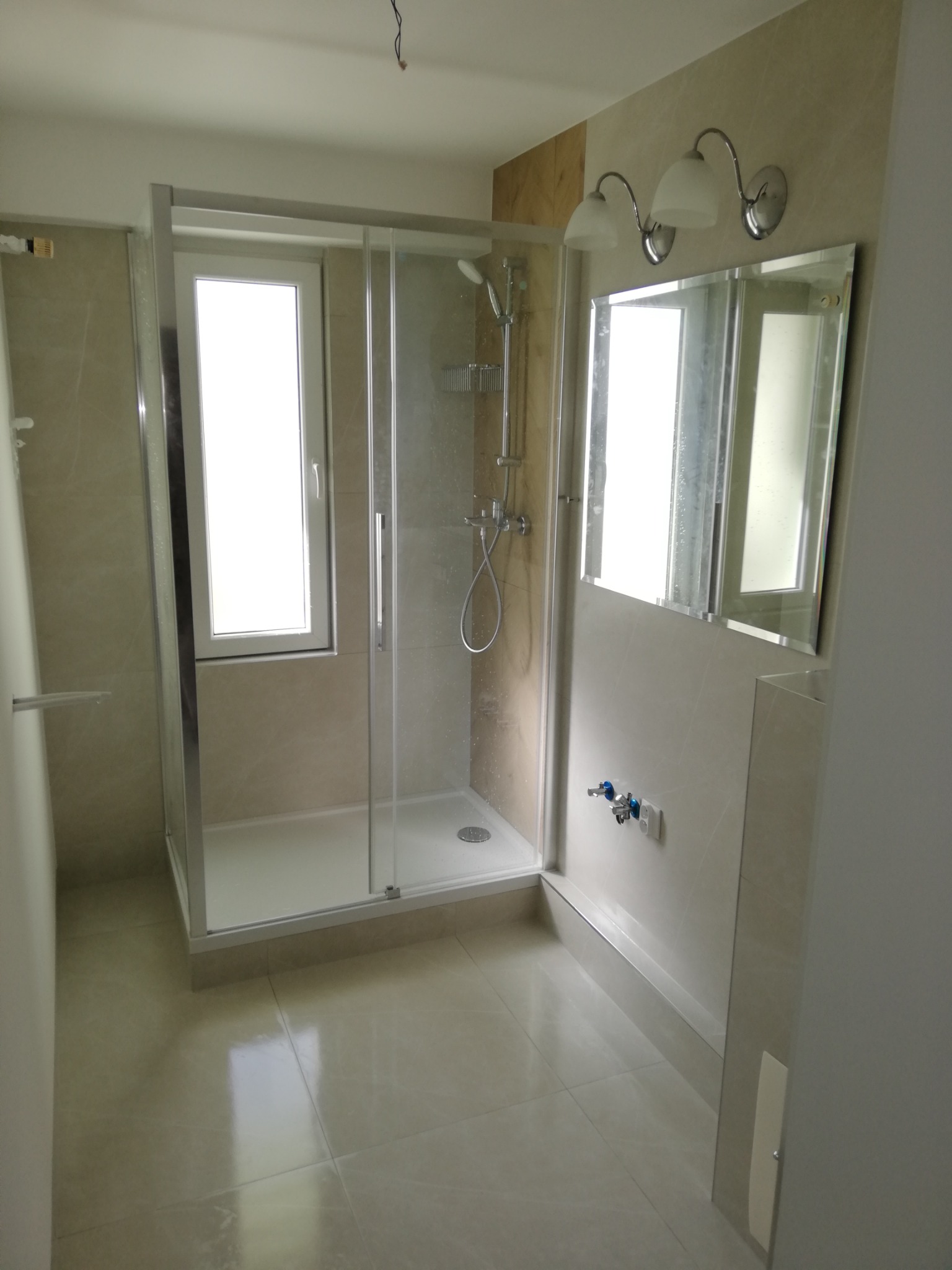 Nowoczesna łazienka po remoncie z kabiną prysznicową z chromowanymi profilami, dużym lustrem i beżowymi płytkami imitującymi marmur.