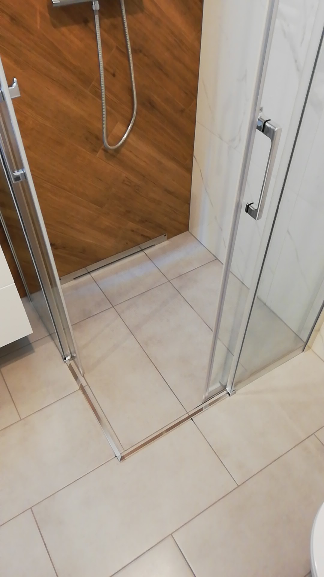 Nowoczesna kabina prysznicowa z chromowanymi profilami, drewnopodobną ścianą i marmurowymi płytkami, widok z góry na beżowe płytki podłogowe.