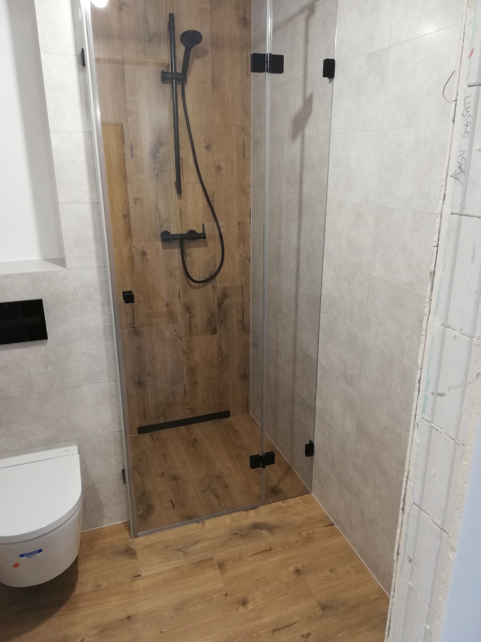 Wykończona łazienka z kabiną prysznicową typu walk-in z czarnymi okuciami, drewnopodobnymi płytkami w strefie prysznica oraz wiszącą toaletą w minimalistycznym stylu.