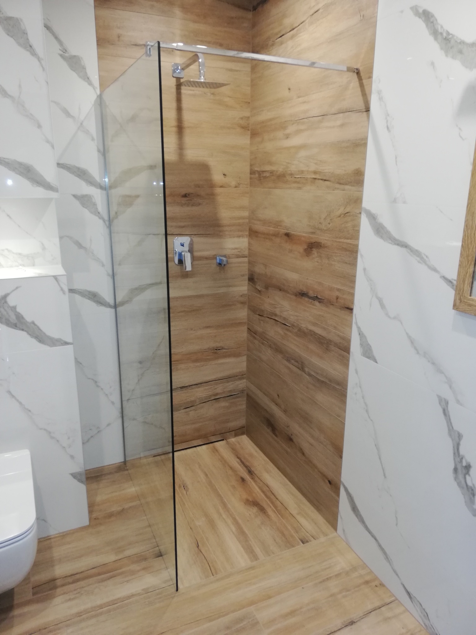 Wykończona łazienka z kabiną prysznicową typu walk-in, wyłożona płytkami imitującymi drewno i marmur, armatura chromowana, widoczny fragment toalety.