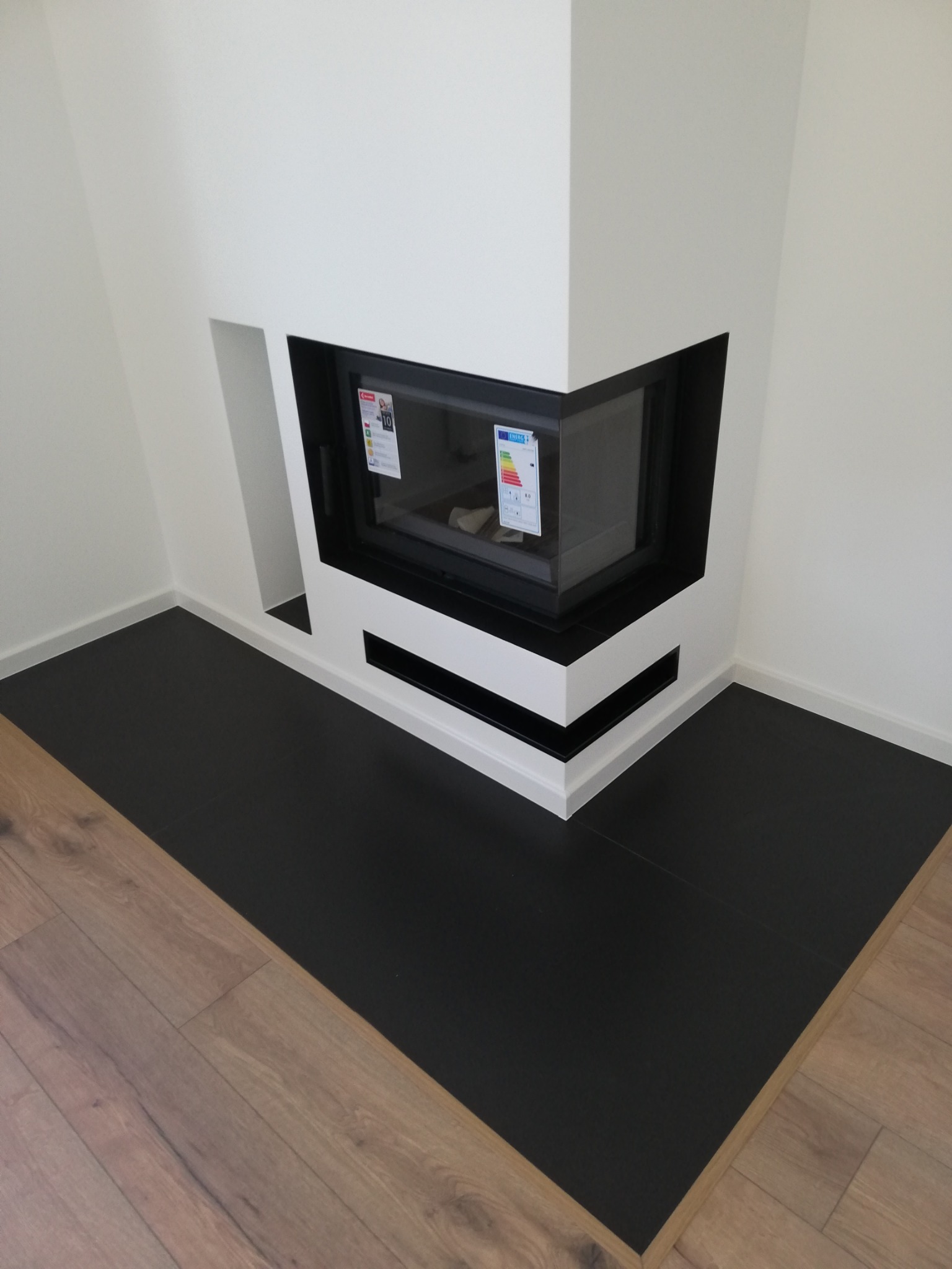 Nowoczesny narożny kominek z czarnym wkładem i białym obramowaniem, widoczne naklejki informacyjne na szybie, czarna podłoga z płytek kontrastująca z drewnianą podłogą w jodełkę.