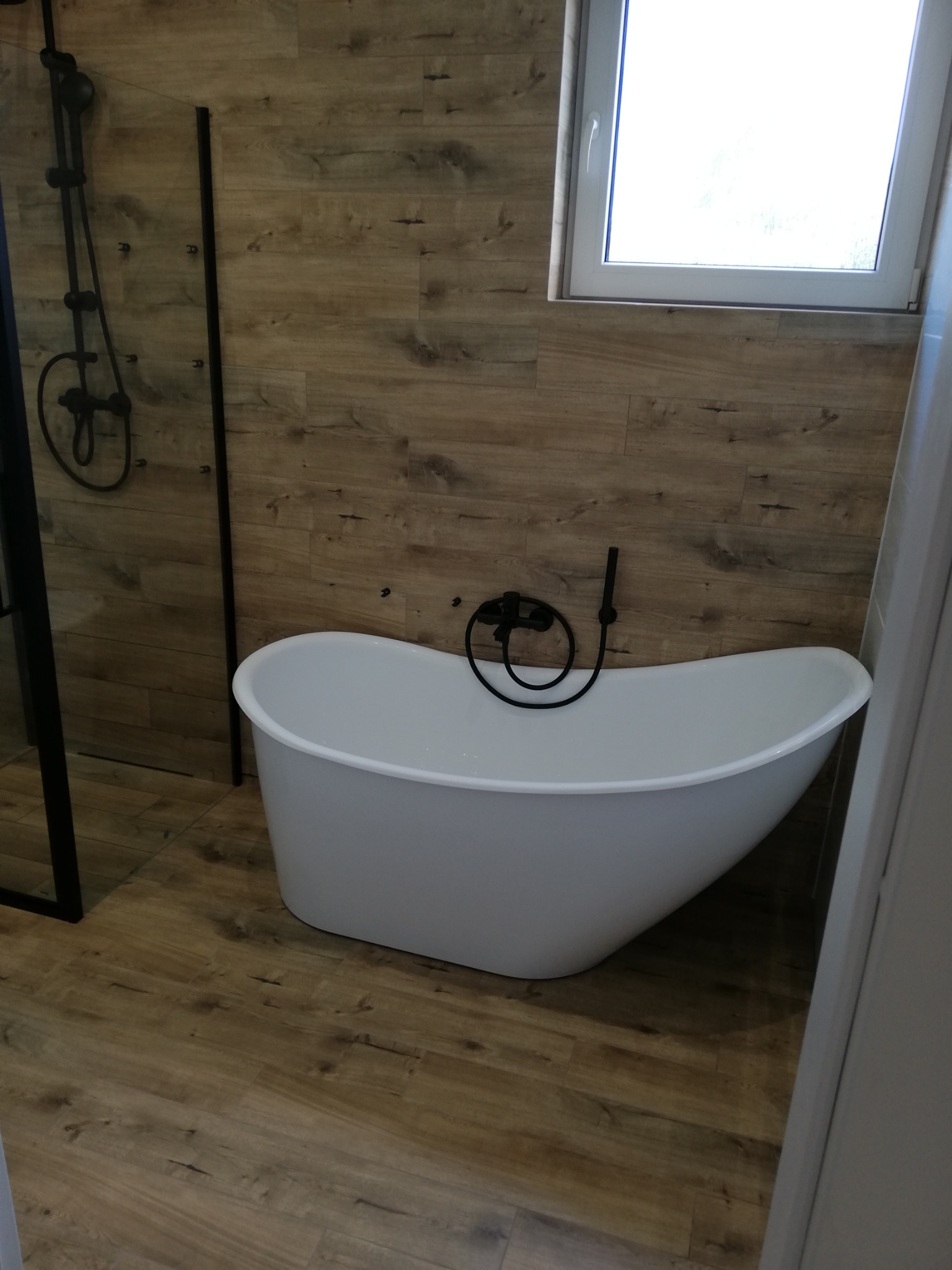 Nowoczesna łazienka z wolnostojącą białą wanną, czarną armaturą i drewnopodobnymi płytkami na ścianach i podłodze, oświetlona światłem dziennym z okna.