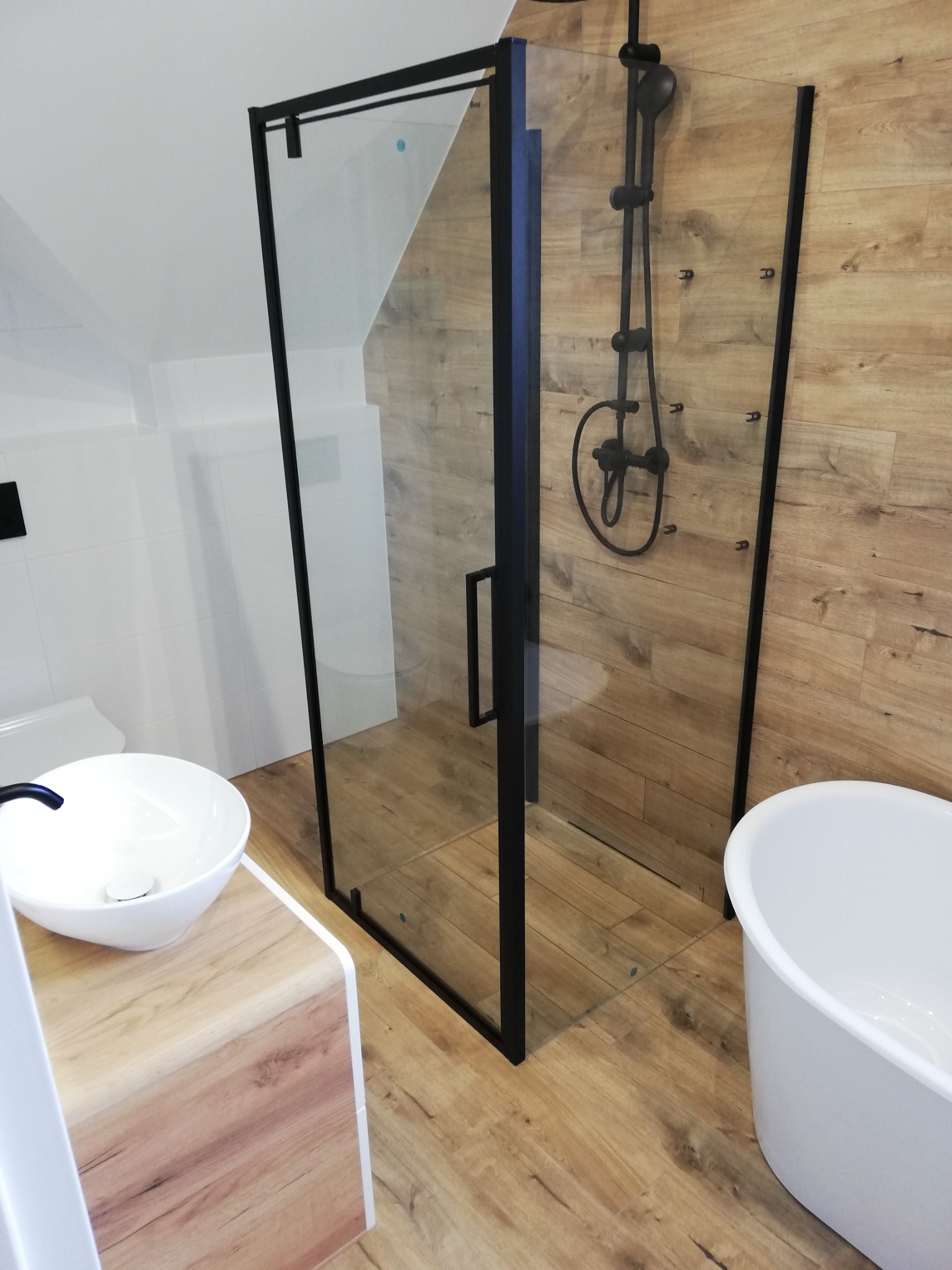 Nowoczesna łazienka z kabiną prysznicową typu walk-in z czarną armaturą, umywalką nablatową i wolnostojącą wanną, wykończona płytkami imitującymi drewno.