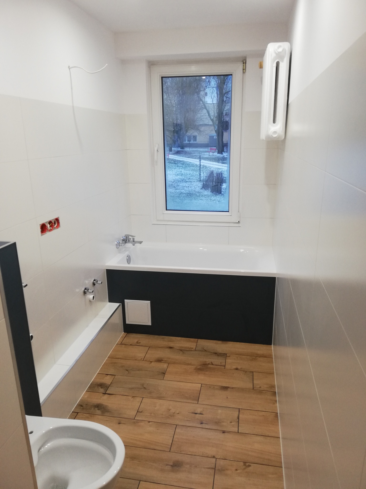 Wykończona łazienka w Olsztynie z wanną obudowaną czarną płytą, drewnopodobną podłogą i białymi płytkami ściennymi, widoczne przygotowane punkty elektryczne i grzejnik.