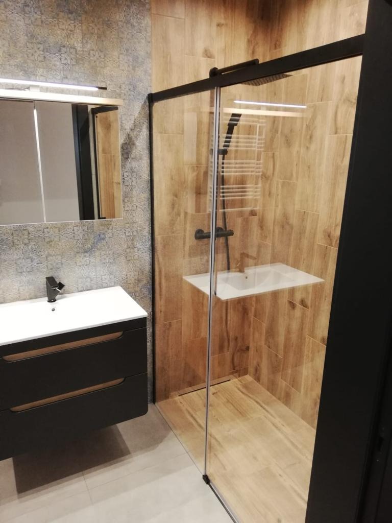 Nowoczesna łazienka z kabiną prysznicową typu walk-in z czarnymi profilami, umywalką nablatową na czarnej szafce i lustrem z oświetleniem LED.