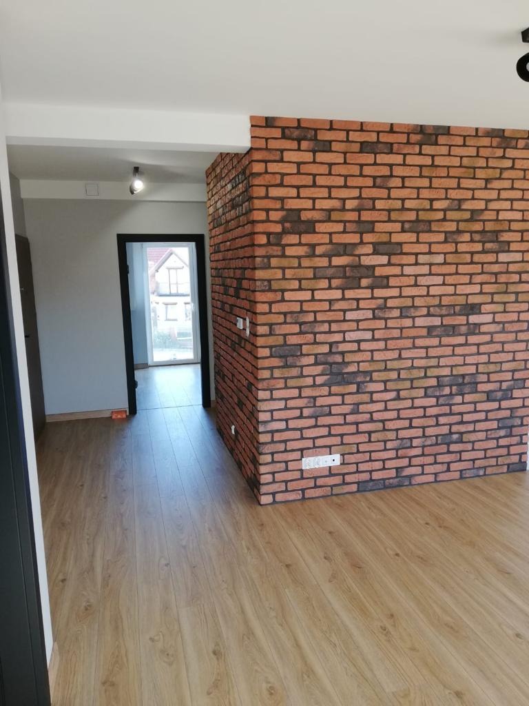 Wnętrze mieszkania z wykończoną ścianą z cegły klinkierowej, widoczna drewnopodobna podłoga i otwarte przejście do kolejnego pomieszczenia z ościeżnicą w kolorze antracytowym.