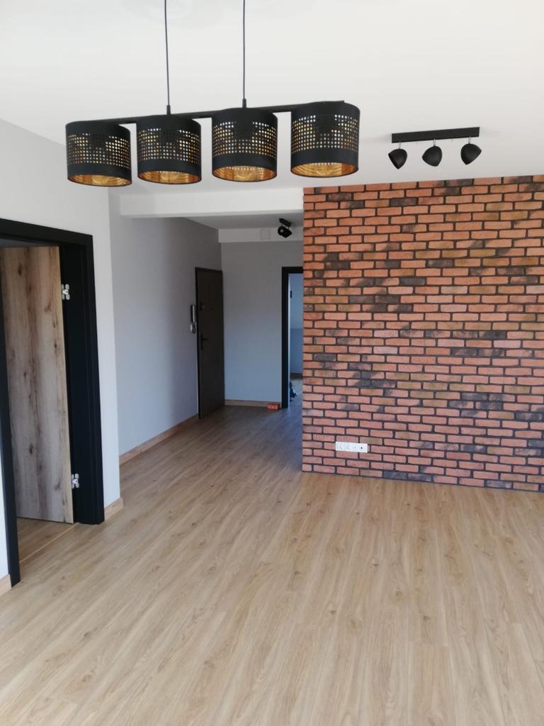 Nowoczesne wnętrze po remoncie: parkiet laminowany w odcieniach beżu, ściana z czerwonej cegły, czarne lampy sufitowe z ażurowymi kloszami i czarne drzwi z drewnianym fornirem.
