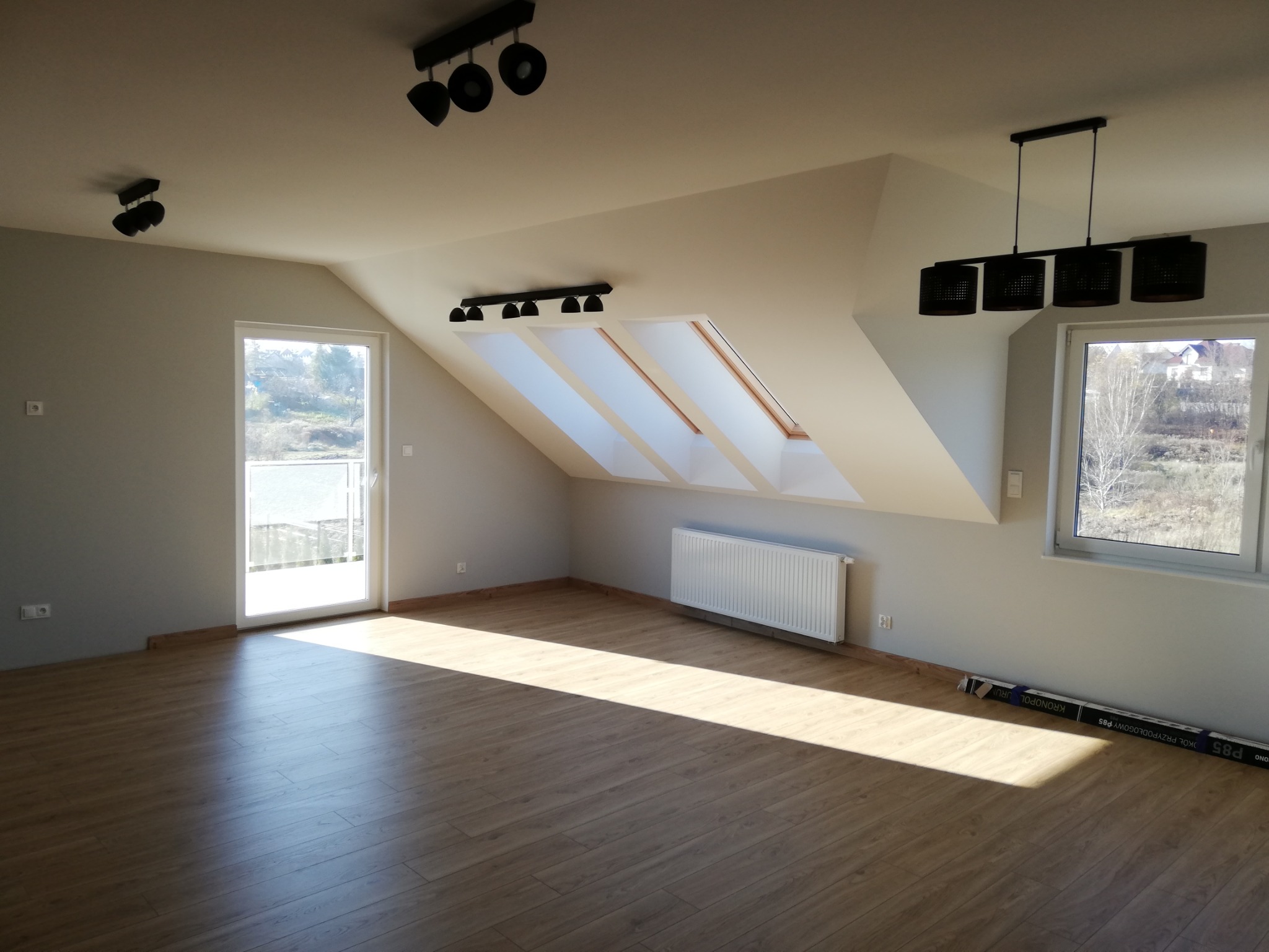 Poddasze po remoncie z drewnopodobną podłogą, trzema oknami dachowymi, grzejnikiem, lampami sufitowymi i listwami przypodłogowymi. Na podłodze leżą opakowania po panelach podłogowych.
