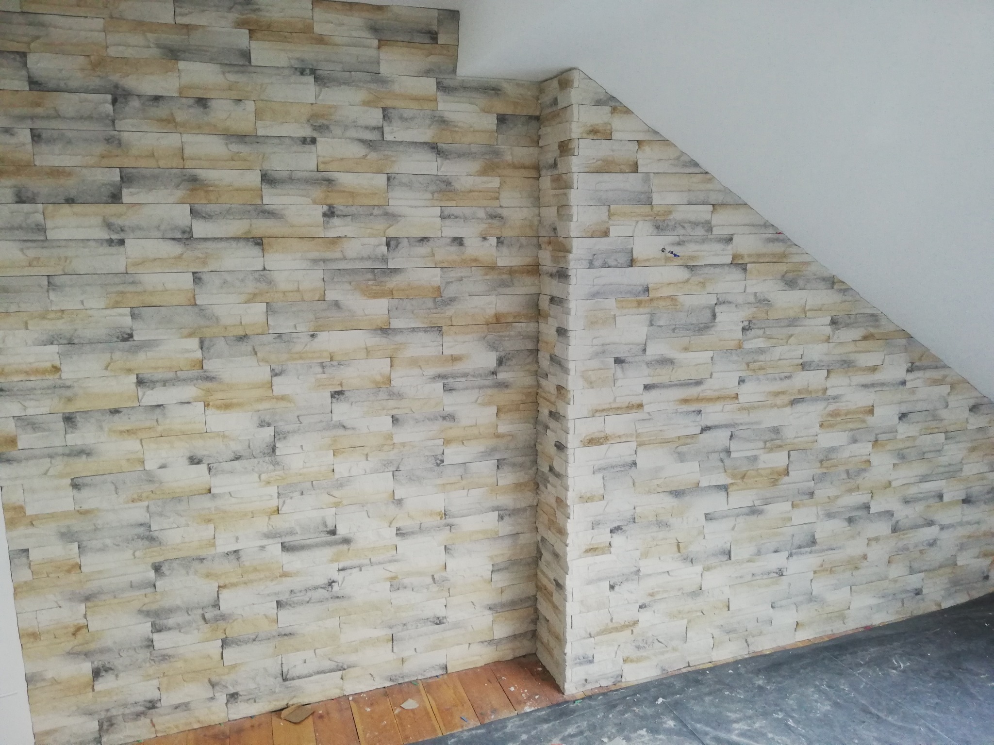 Ściana wykończona dekoracyjną okładziną imitującą kamień w odcieniach beżu, szarości i bieli, w rogu pomieszczenia na poddaszu, gdzie łączy się ze ścianą pomalowaną na biało. Część podłogi...