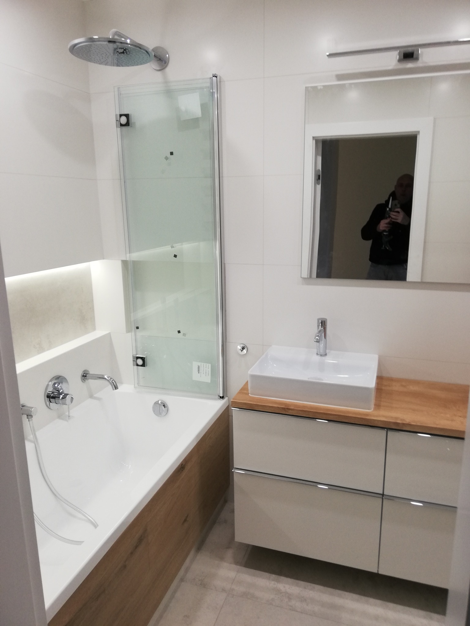 Nowoczesna łazienka z białą armaturą, kabiną prysznicową ze składanym szklanym panelem, umywalką nablatową na szafce z drewnianym blatem i lustrem, w którym odbija się osoba robiąca zdjęcie.