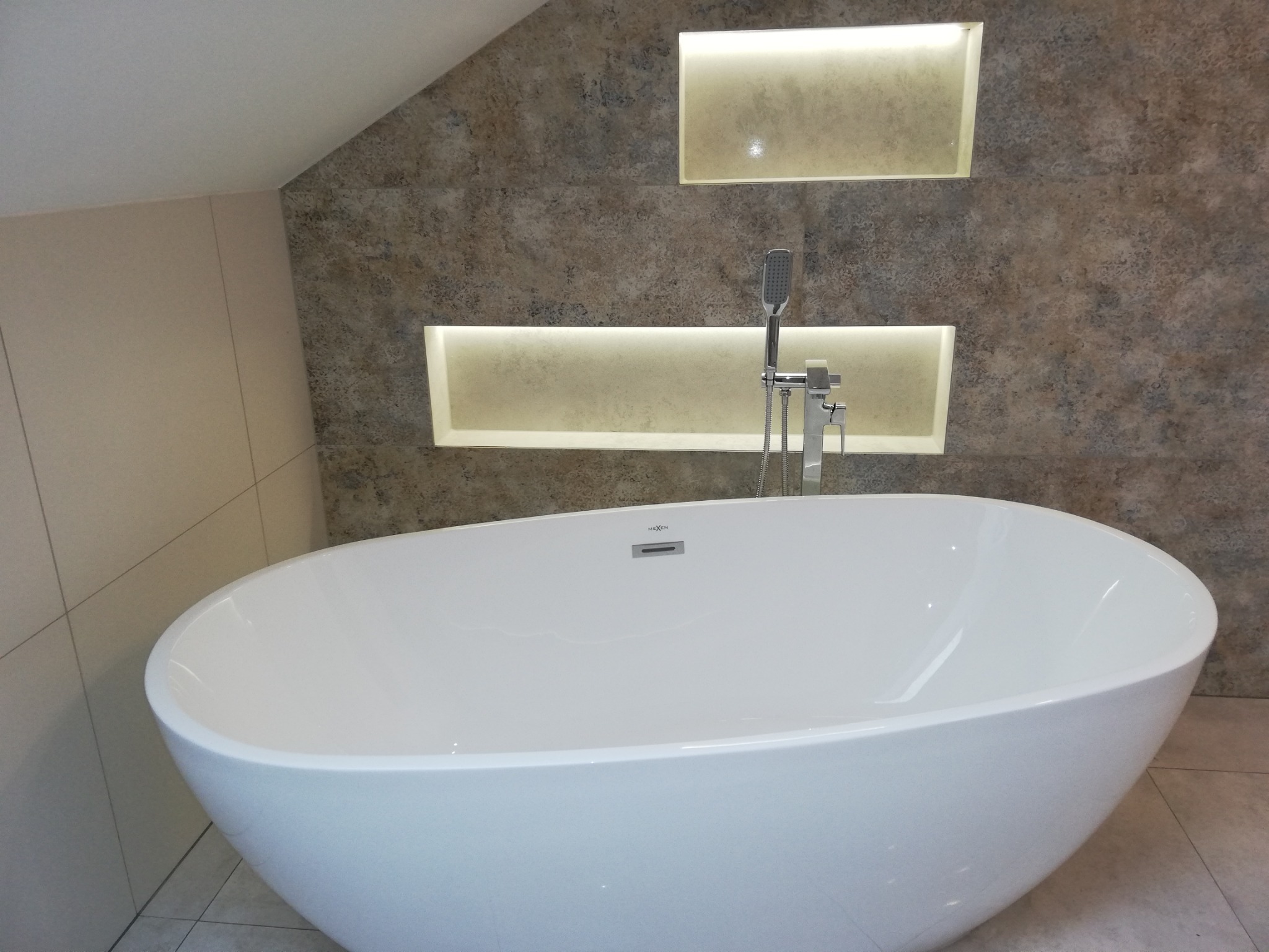 Biała, wolnostojąca wanna owalna z chromowaną baterią i słuchawką prysznicową na tle ściany z dekoracyjnymi, podświetlanymi wnękami w wykończonej łazience na poddaszu.