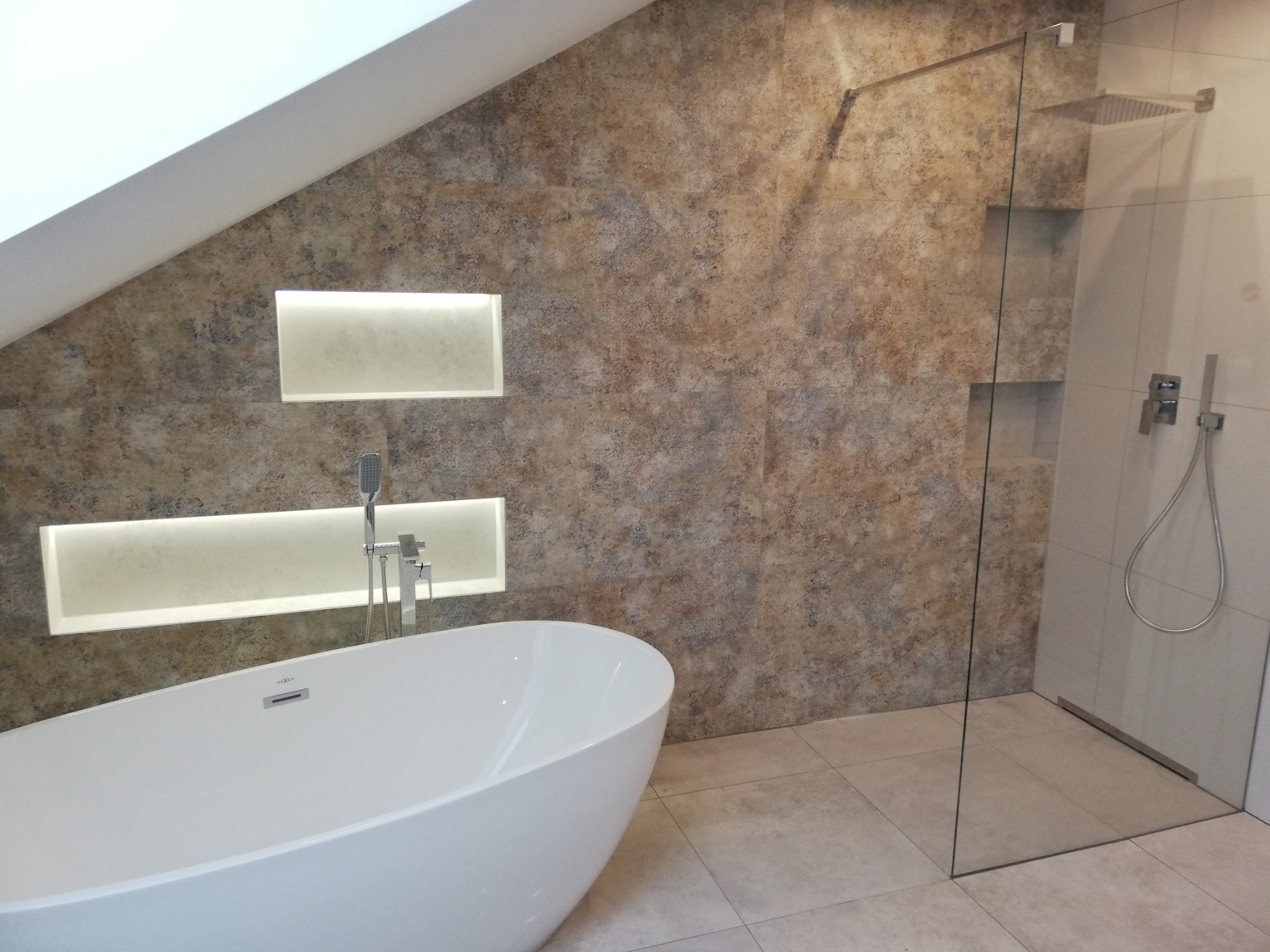 Nowoczesna łazienka na poddaszu z wolnostojącą białą wanną, kabiną prysznicową typu walk-in z deszczownicą i podświetlanymi wnękami w ścianie wyłożonej płytkami imitującymi kamień.