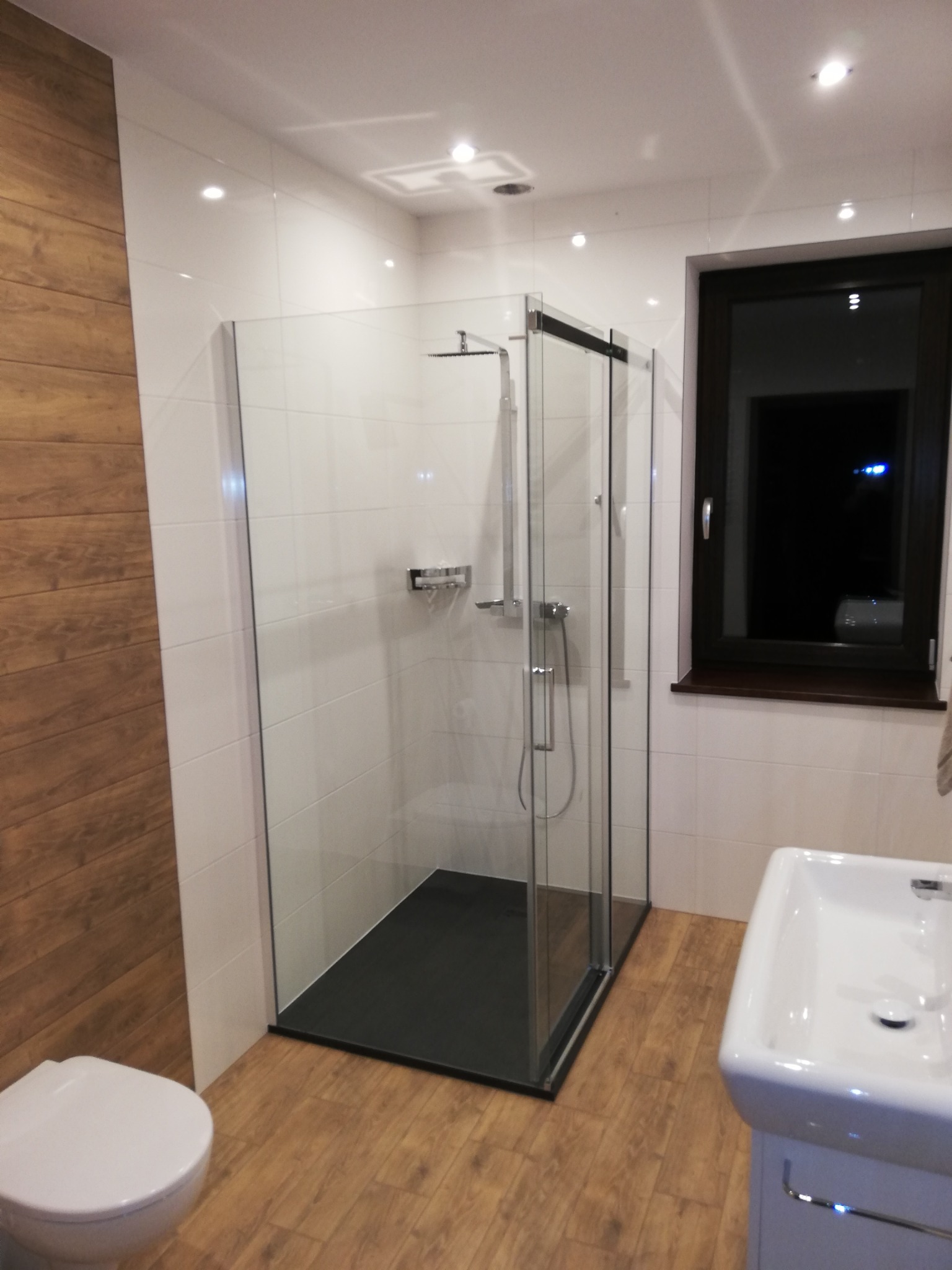 Nowoczesna łazienka z kabiną prysznicową typu walk-in z czarnym brodzikiem, białymi płytkami i drewnopodobną okładziną ściany. Widoczna również biała umywalka i toaleta.