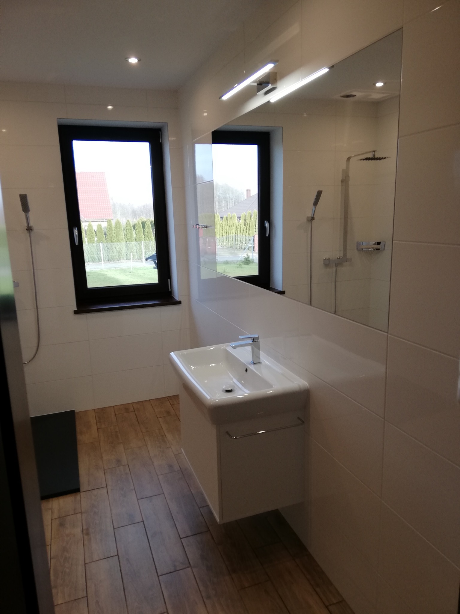 Nowoczesna łazienka z białymi płytkami, drewnopodobną podłogą i wiszącą umywalką z szafką, okno z widokiem na ogród odbija się w lustrze.