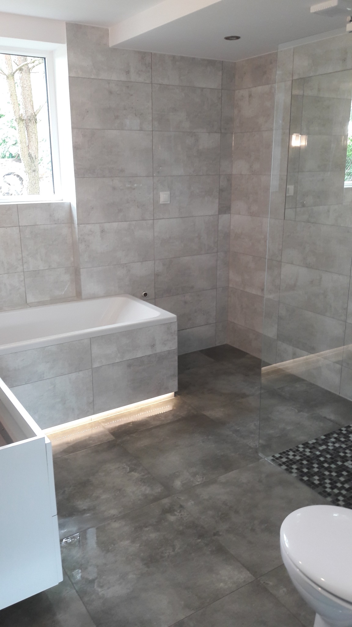 Nowoczesna łazienka z szarymi płytkami imitującymi kamień, podświetlana wanna, szklana kabina prysznicowa i wisząca toaleta.