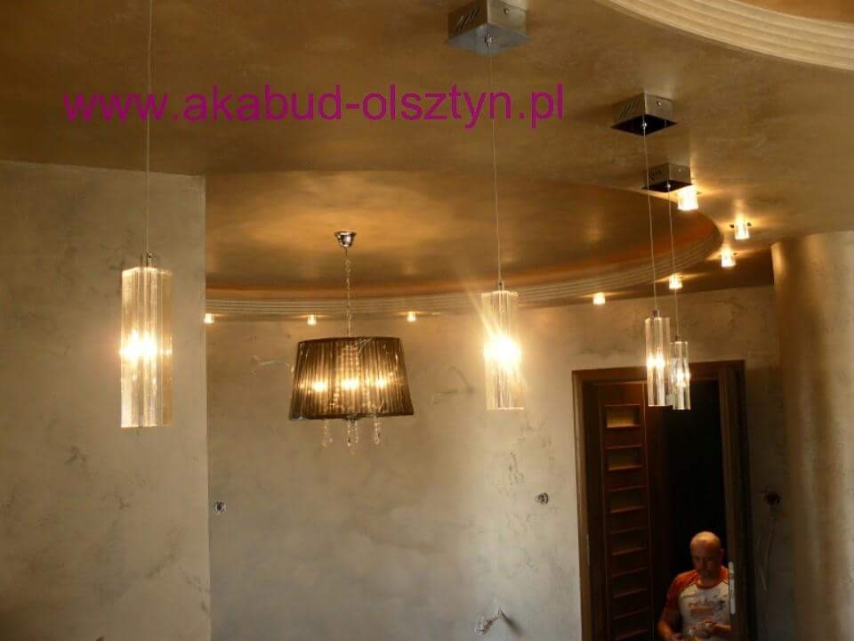 Wykończone wnętrze z dekoracyjną, metalizowaną ścianą i sufitem, oświetlone wiszącymi lampami z kryształkami, widoczny fragment otwartych drzwi i sylwetka mężczyzny.