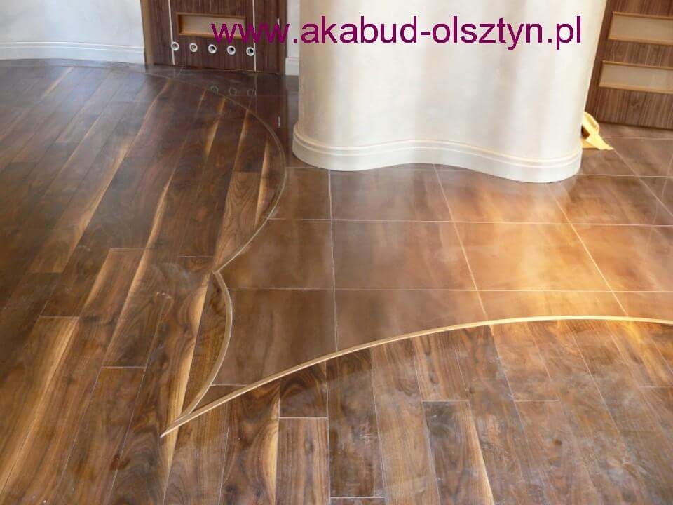 Parkiet z ciemnego drewna ułożony we wzór z jasnymi intarsjami, przechodzący w kwadratowe płytki, widoczny fragment drzwi i kolumny.