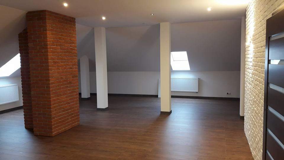 Poddasze po remoncie z drewnopodobną podłogą, ceglanym filarem i białymi ścianami, oświetlone światłem z okien dachowych.