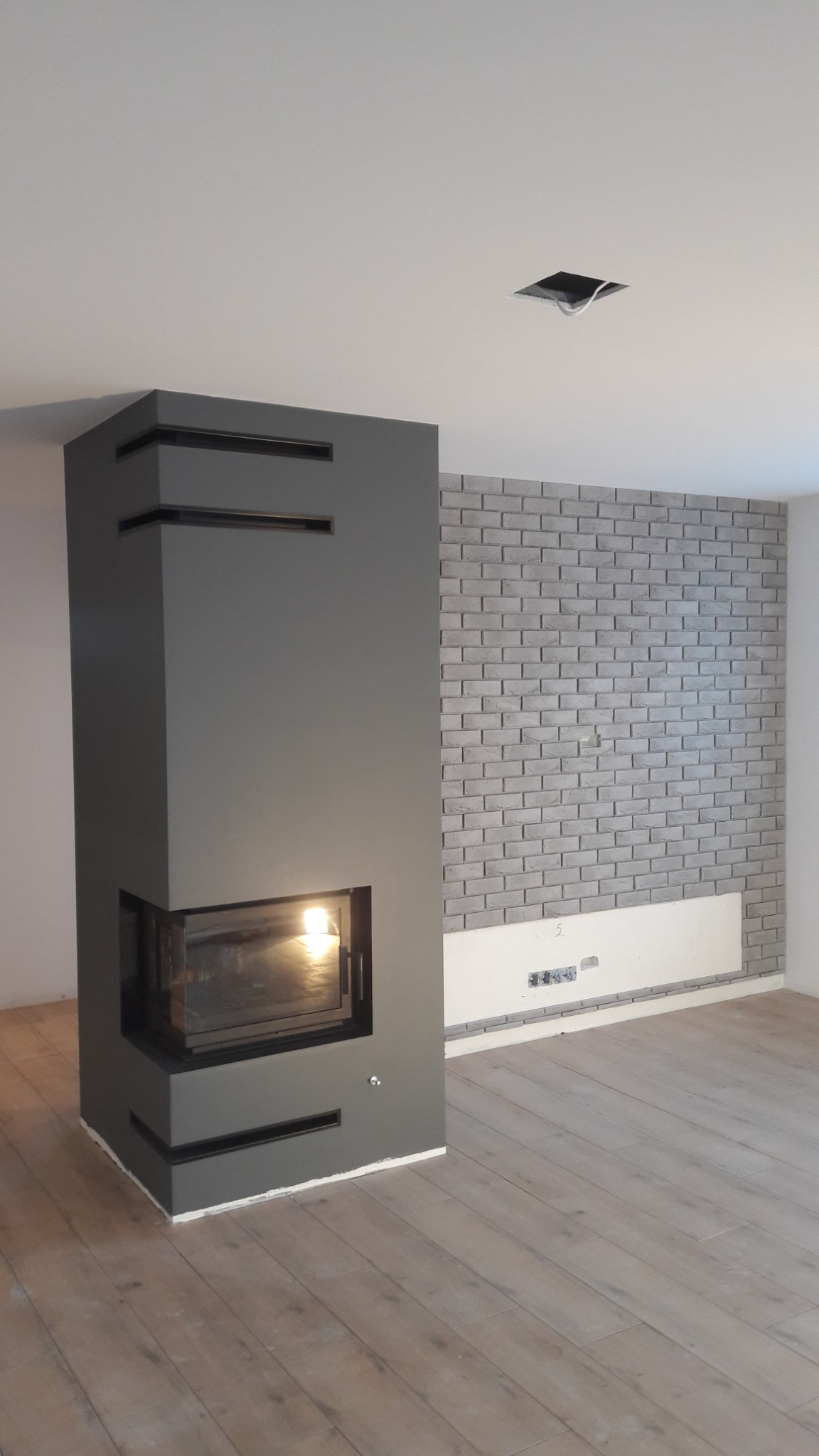 Nowoczesny kominek obudowany szarą konstrukcją z poziomymi wnękami, z palącym się ogniem wewnątrz, na tle ściany wyłożonej szarymi cegłami w nowym wnętrzu z drewnianą podłogą.