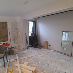 Malmaster - Montaż okna w surowym wnętrzu: pracownik na kolanach uszczelnia okno, widoczne narzędzia, drabina i deski na podłodze, ściany przygotowane do wykończenia.