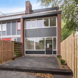 Malmaster - Nowoczesny dom szeregowy z elewacją z szarej blachy falistej i cegły, otoczony drewnianym płotem i kostką brukową na podjeździe.