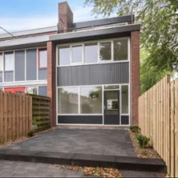 Nowoczesny dom szeregowy z elewacją z szarej blachy falistej i cegły, otoczony drewnianym płotem i kostką brukową na podjeździe.