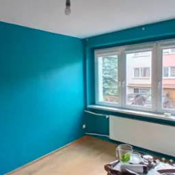 Pomieszczenie po malowaniu ścian na turkusowo, widoczne okno z widokiem na zewnątrz i grzejnik. Na podłodze widoczne akcesoria malarskie.
