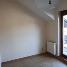 Jasne wnętrze pokoju na poddaszu z drewnianą podłogą, beżowymi ścianami, białym grzejnikiem i oknem balkonowym z widokiem na zewnątrz. Minimalistyczny design.