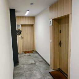Malmaster - Korytarz w bloku z szarymi płytkami podłogowymi i drewnopodobnymi drzwiami do mieszkań nr 9 i 10, z wycieraczkami i wieńcem na drzwiach.