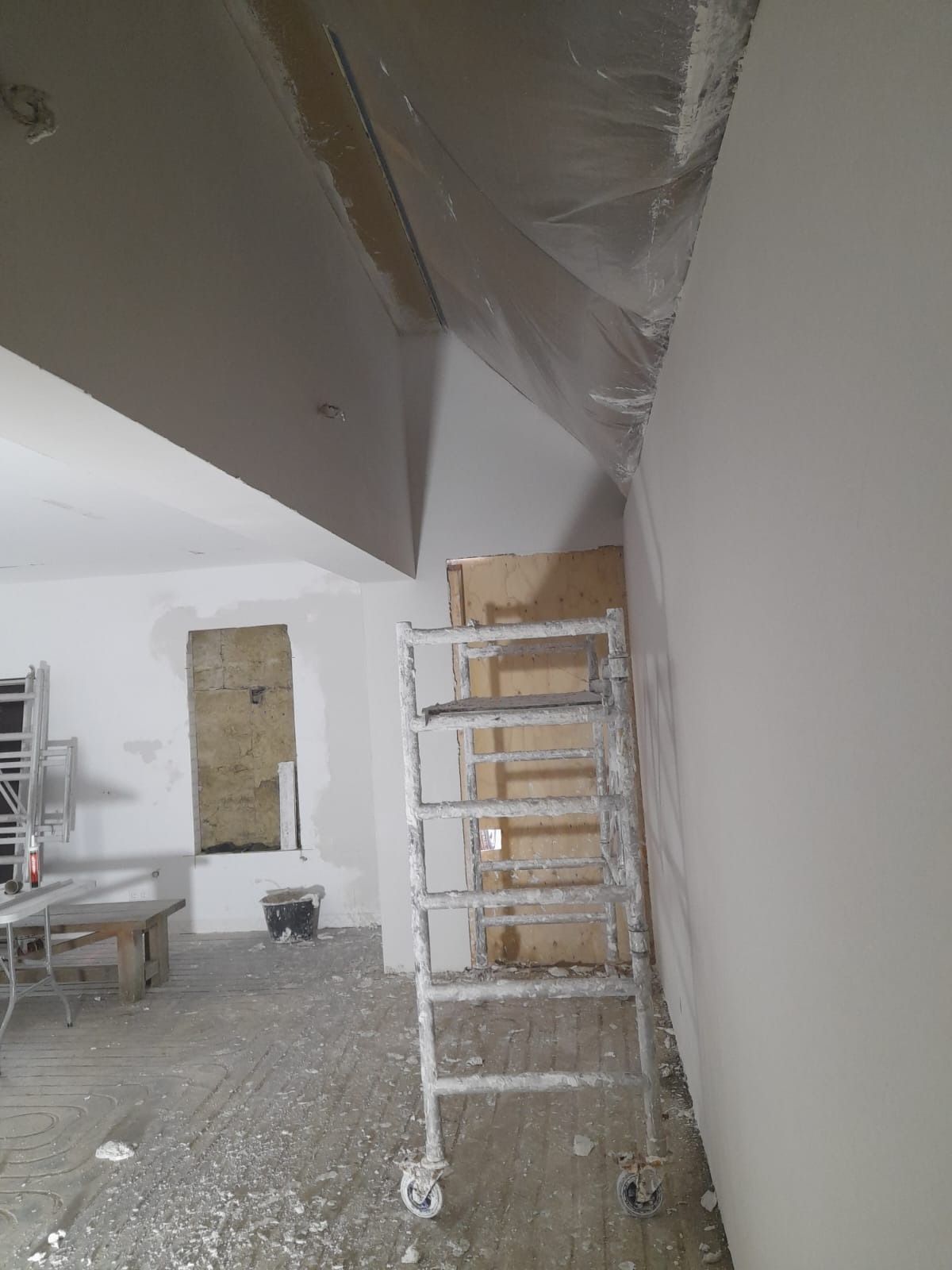 Pokój w trakcie szpachlowania i malowania, widoczna drabina malarska, zakryte folią zabezpieczającą fragmenty sufitu, ślady gładzi na podłodze i ścianach, oraz niezabezpieczone okno.