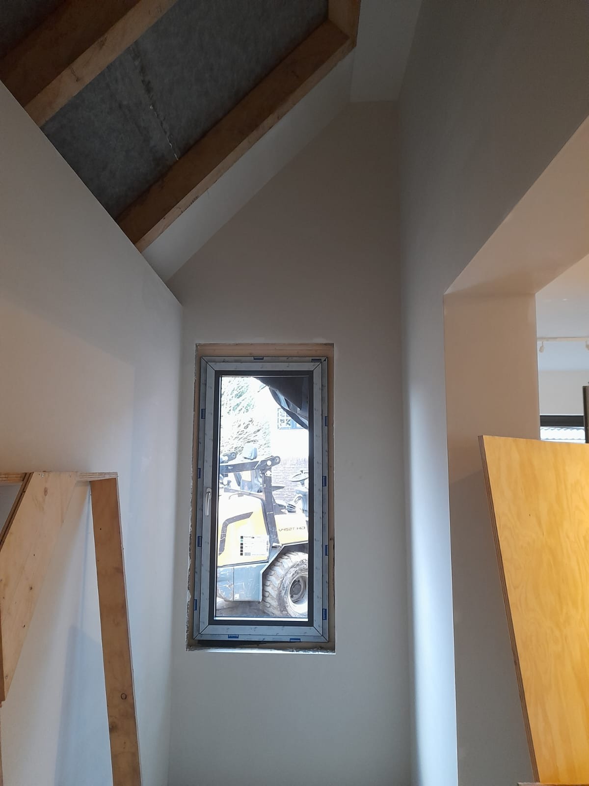 Wnętrze w trakcie prac wykończeniowych z widoczną surową konstrukcją dachu, świeżo oszlifowanymi ścianami i oknem z widokiem na maszynę budowlaną na zewnątrz.