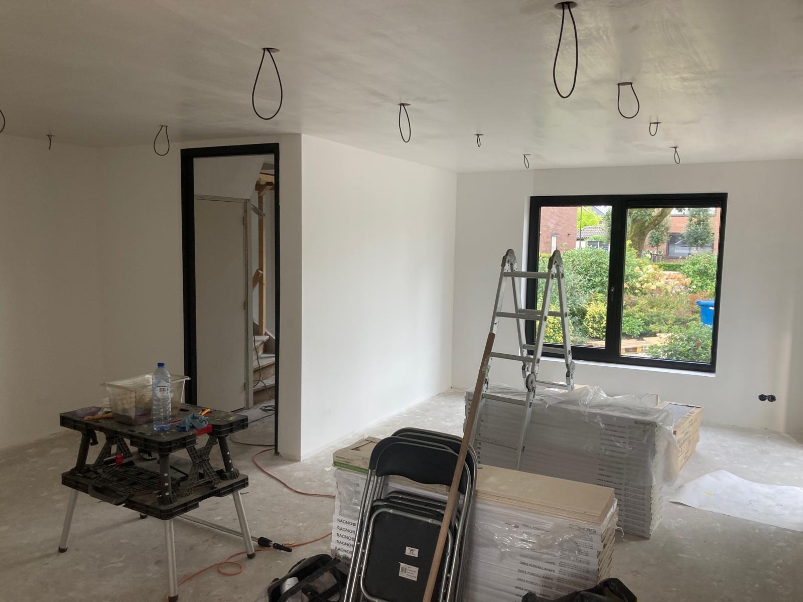 Wnętrze w Rościsławicach w trakcie prac wykończeniowych: ściany po szpachlowaniu, przygotowane do malowania, widoczne kable oświetleniowe, drabina oparta o okno, ułożone płytki podłogowe, narzędzia...