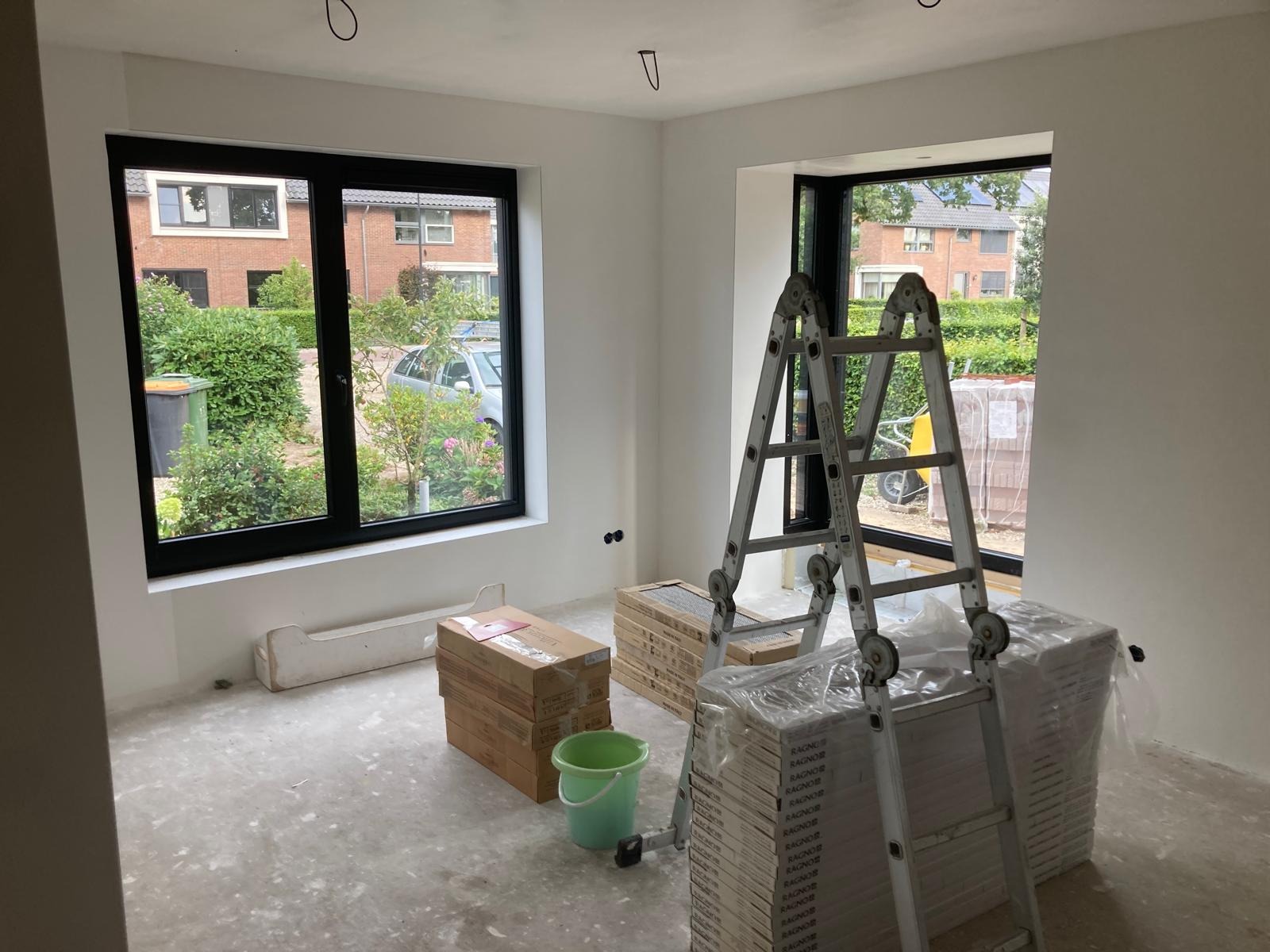Przygotowania do prac wykończeniowych w jasnym pokoju z dwoma oknami w czarnych ramach, widoczne kartony z panelami, drabina aluminiowa, ślady po szpachlowaniu na podłodze i ścianach.