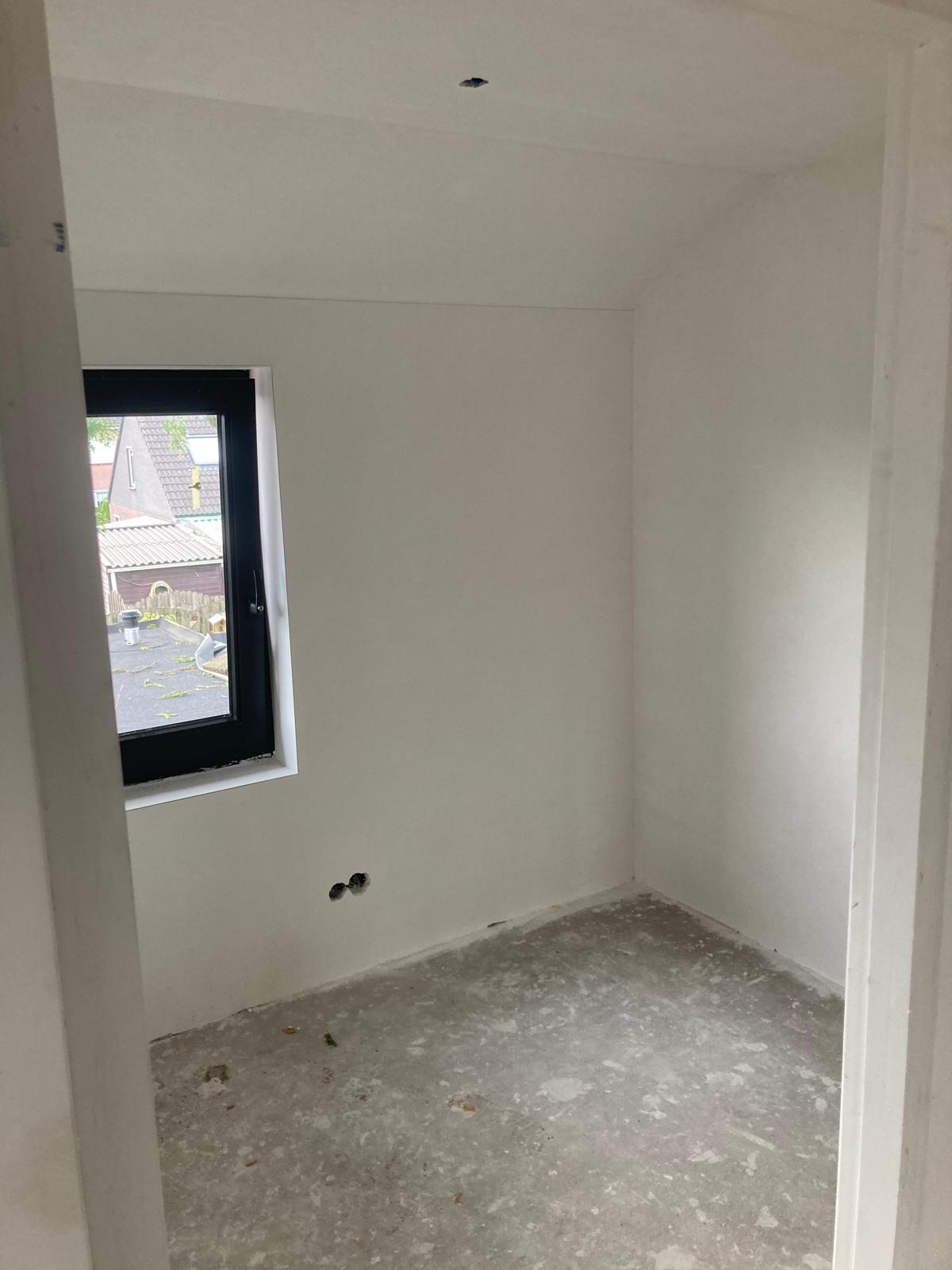 Pusty pokój po szpachlowaniu i malowaniu na biało, z widocznymi gniazdkami elektrycznymi i czarną ramą okna, surowy beton na podłodze.