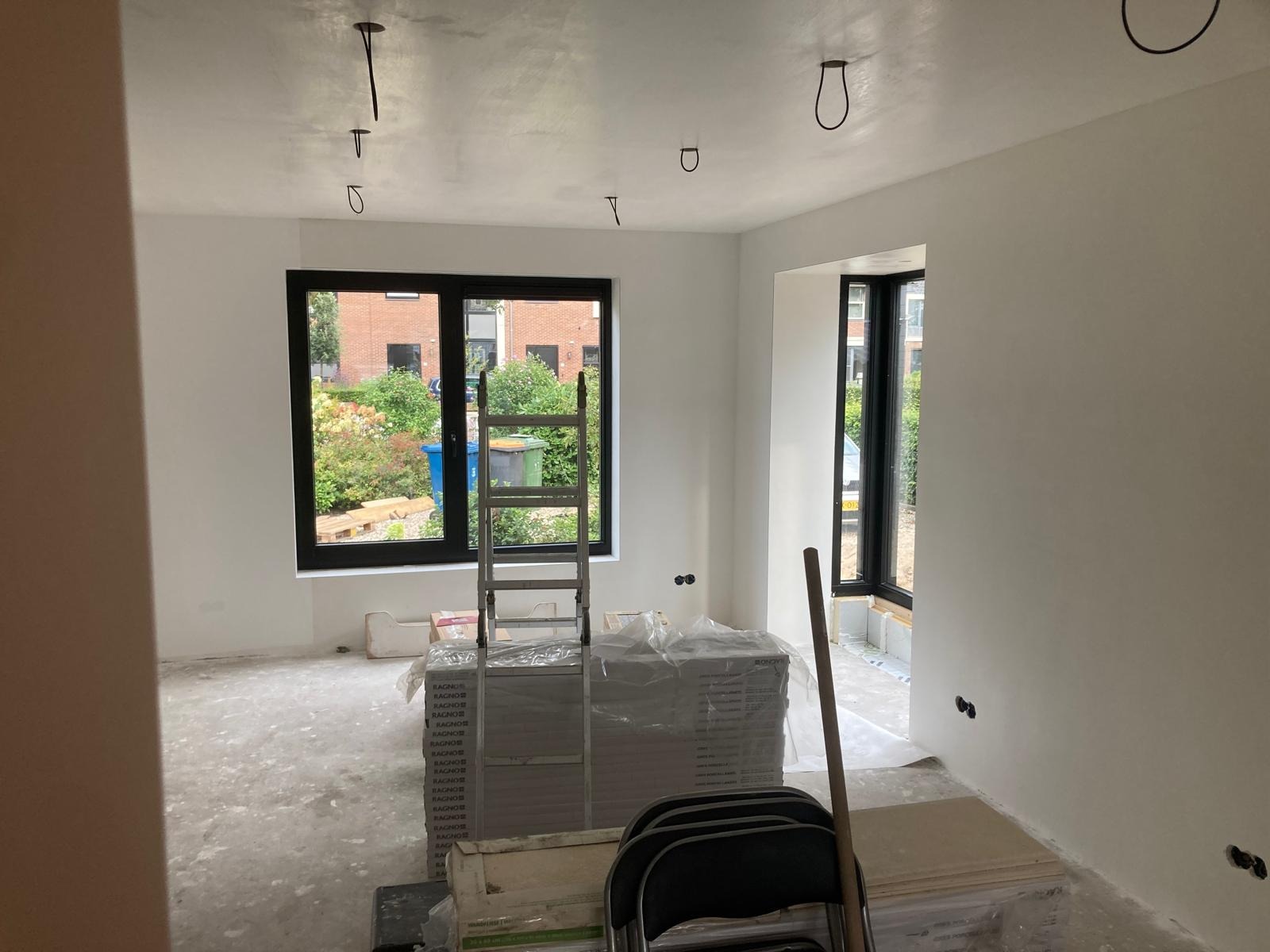 Wnętrze w trakcie prac wykończeniowych: białe ściany, surowy beton na podłodze, okna z czarnymi ramami, drabina oparta o okno, przygotowane płyty podłogowe i widoczne punkty elektryczne.