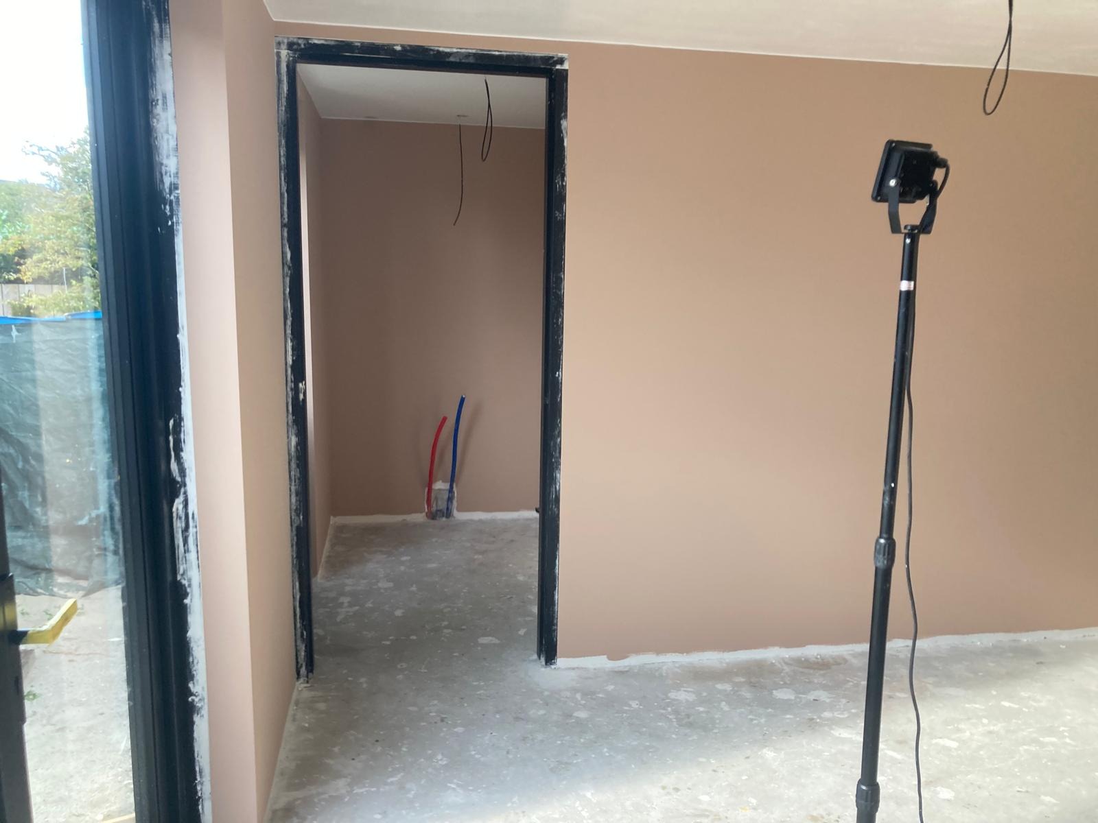 Pokój po szpachlowaniu ścian na gładko, widoczne rury instalacji wodnej wystające ze ściany, czarne futryny drzwiowe i lampa oświetleniowa na statywie.