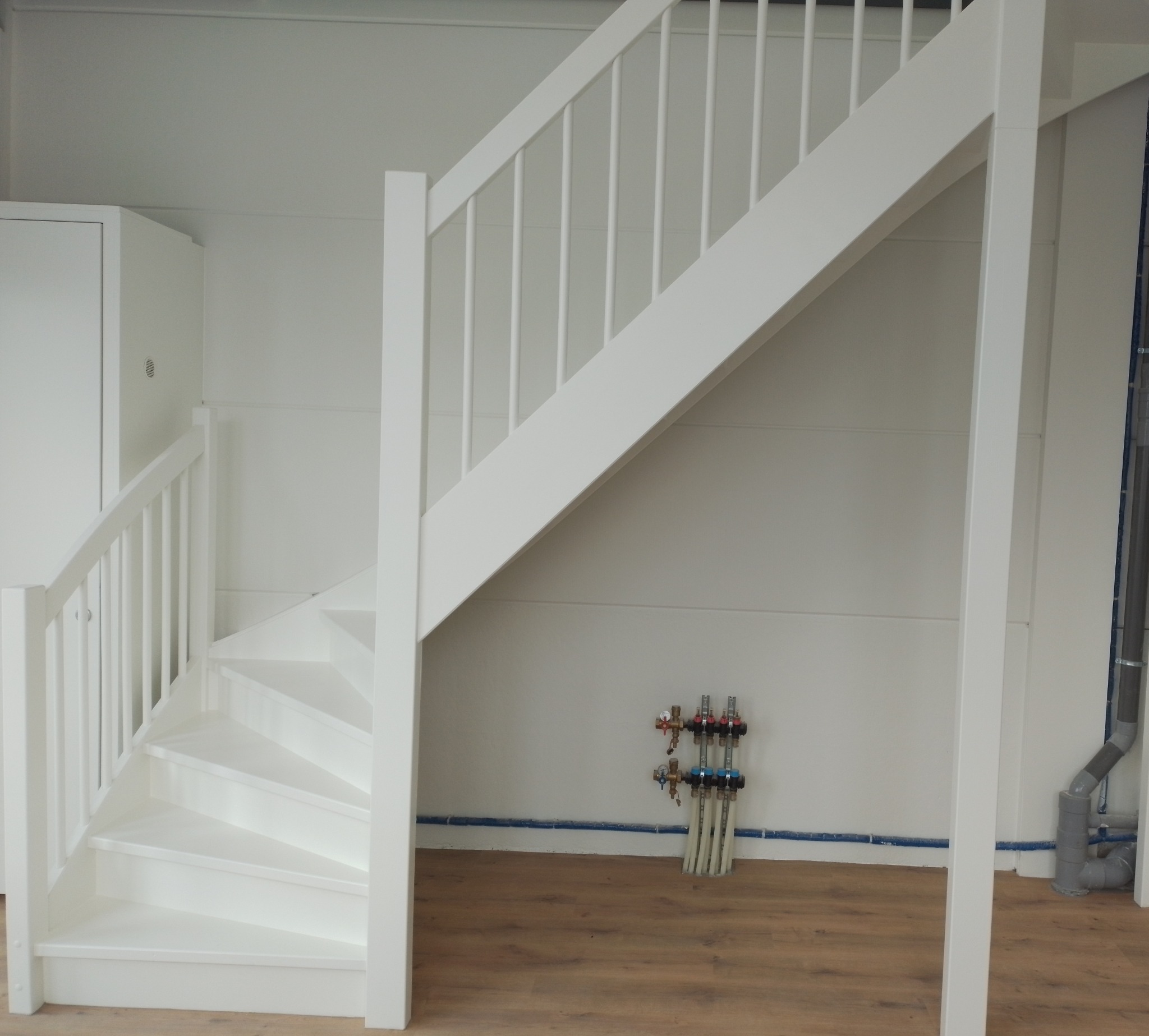 Białe, drewniane schody w jasnym pomieszczeniu z panelami podłogowymi, widoczne instalacje hydrauliczne przy ścianie.