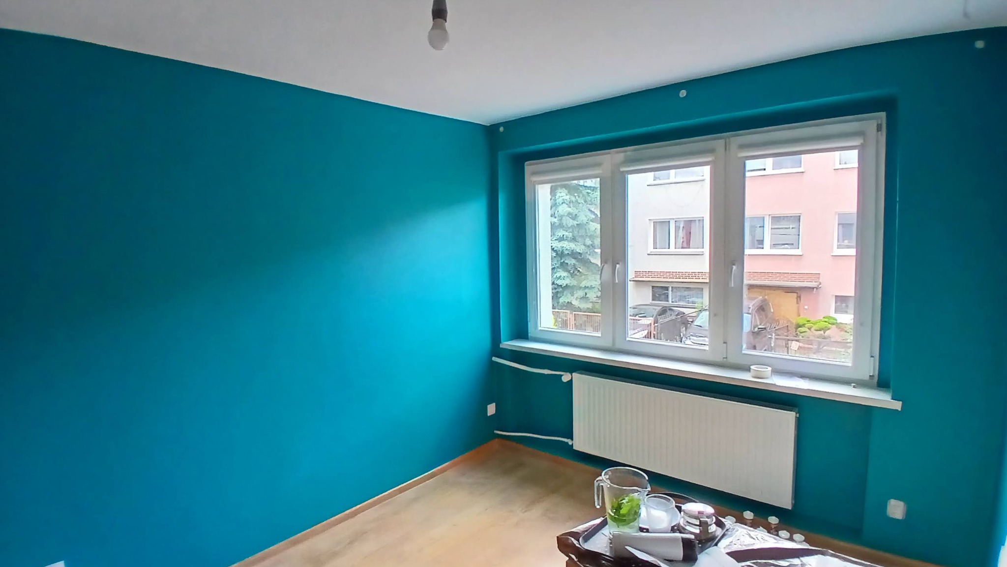 Pomieszczenie po malowaniu ścian na turkusowo, widoczne okno z widokiem na zewnątrz i grzejnik. Na podłodze widoczne akcesoria malarskie.