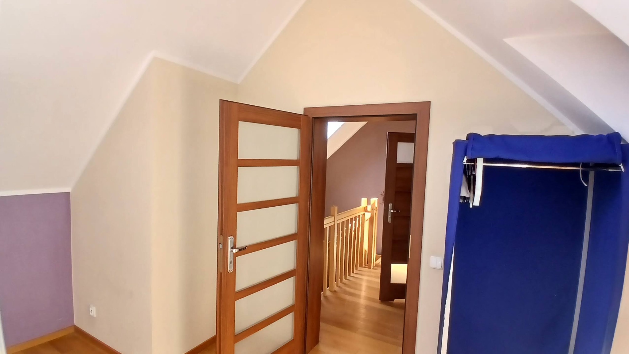 Poddasze po remoncie: drewniane drzwi z przeszkleniami, widok na schody z drewnianą balustradą, ściany w odcieniach beżu i fioletu, garderoba przenośna w kolorze niebieskim.