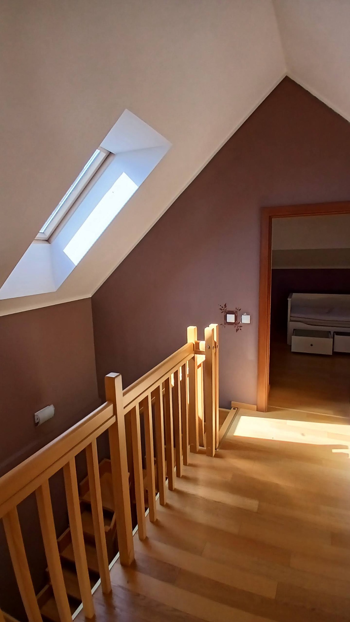 Poddasze z drewnianą balustradą schodów i brązową ścianą z oknem dachowym. Widoczna podłoga z paneli i fragment pokoju z białymi meblami w tle.