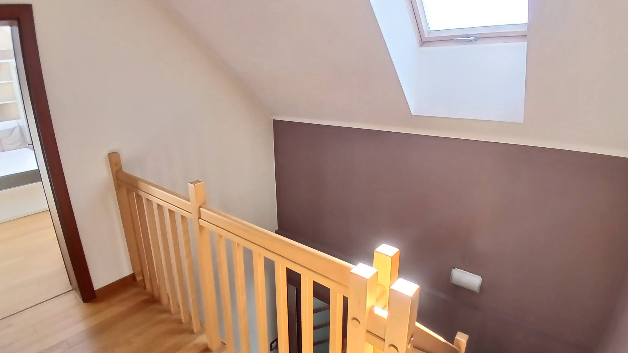 Jasne drewniane schody z barierką w domu z brązową ścianą i oknem dachowym, widok z góry. Częściowo widoczny pokój z białymi meblami.