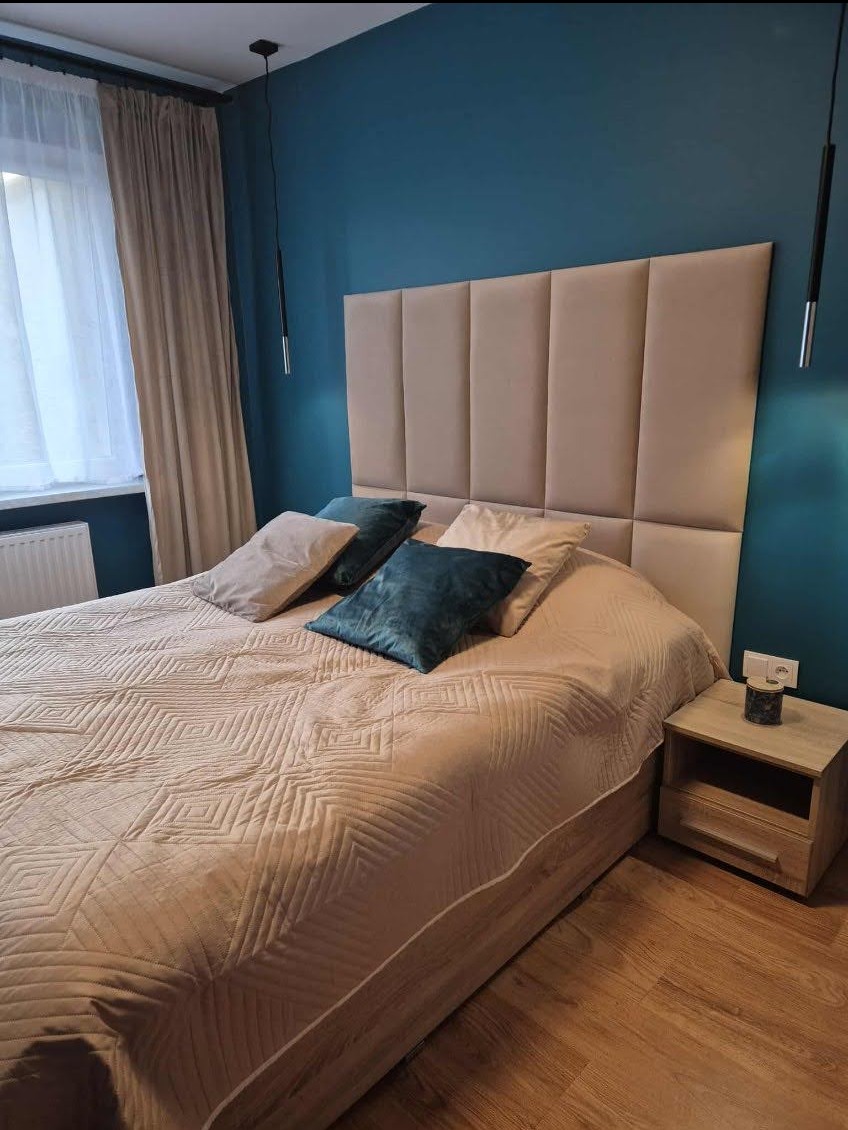 Wykończenie sypialni w Rościsławicach: łóżko z beżowym zagłówkiem i narzutą w geometryczne wzory, na drewnianej podłodze, z turkusową ścianą w tle.