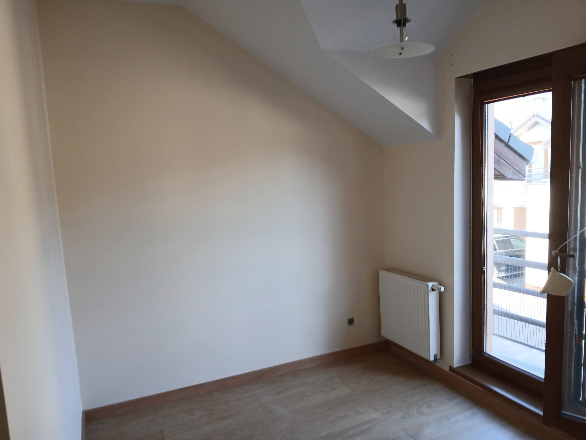 Jasne wnętrze pokoju na poddaszu z drewnianą podłogą, beżowymi ścianami, białym grzejnikiem i oknem balkonowym z widokiem na zewnątrz. Minimalistyczny design.