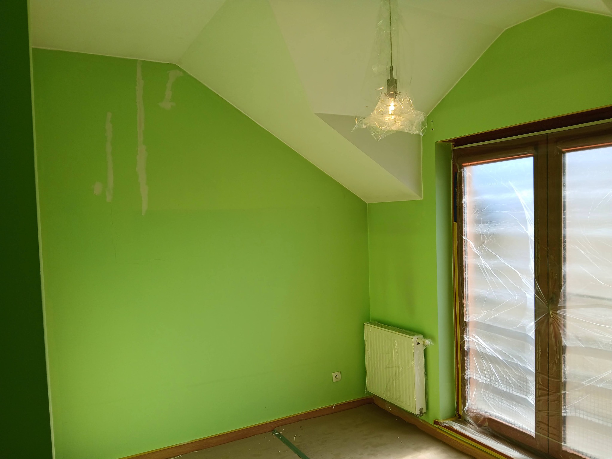 Pokój po szpachlowaniu, przed malowaniem na zielono. Widoczne białe pasy szpachli na ścianie, okno i grzejnik zabezpieczone folią malarską, lampa w folii.